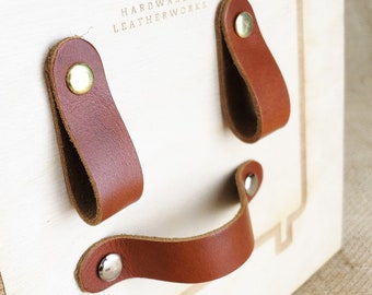 May include: Three brown leather handles with brass and silver rivets mounted on a light wood panel. The panel has the text "Keyway Hardware & Leatherworks" engraved on it.