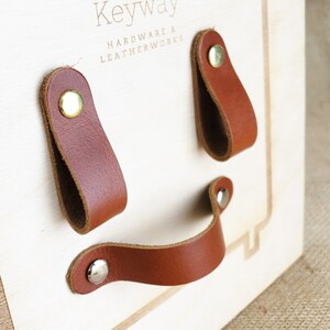 Könnte beinhalten: Drei braune Ledergriffe mit Messing- und Silbernieten, die auf einer hellen Holzplatte montiert sind. Die Platte trägt die eingravierte Aufschrift "Keyway Hardware & Leatherworks".