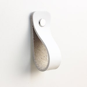 May include: A white leather cabinet pull with a silver rivet. The pull is looped and attached to a white surface. The interior of the loop is a light beige colour.