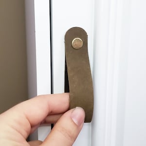 May include: A brown leather cabinet pull with a gold metal screw. The pull is attached to a white cabinet door.