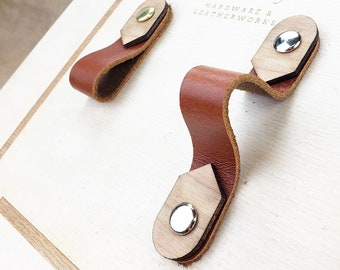 May include: Two leather handles with wooden bases and metal accents. The handles are brown and the bases are light brown. The metal accents are silver and gold. The handles are attached to a white surface with the text "Keyway Hardware & Leatherworks" printed on it.