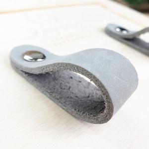 May include: Two gray leather drawer pulls with silver metal accents. The pulls are shaped like a loop and are attached to a wooden surface.