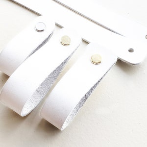 May include: Three white leather drawer pulls with different coloured metal accents. The pulls are all the same size and shape, but have different coloured metal accents. The first pull has a silver accent, the second pull has a gold accent, and the third pull has a brass accent.
