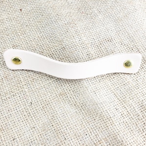 May include: A white leather handle with two gold metal rivets. The handle is curved and has a smooth surface.