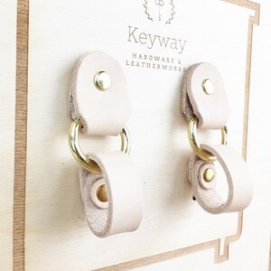 Könnte beinhalten: Zwei hellbraune Lederschlaufen mit Messingringen und Nieten, die auf einem Holzdisplay montiert sind. Das Display trägt den Text "Keyway Hardware & Leatherworks".