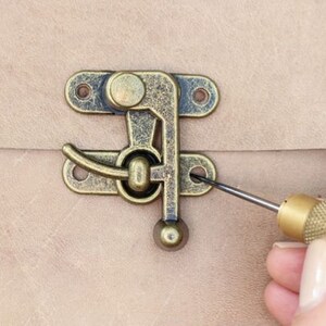 Pode incluir: Um close-up de uma trava de lat&atilde;o sendo fixada com uma pequena chave de fenda. A trava tem um design decorativo e est&aacute; presa a uma superf&iacute;cie de couro marrom claro.