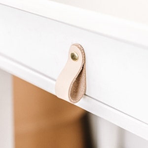 Puede incluir: Mesita de noche blanca con un solo cajón que presenta una manija de cuero marrón claro.