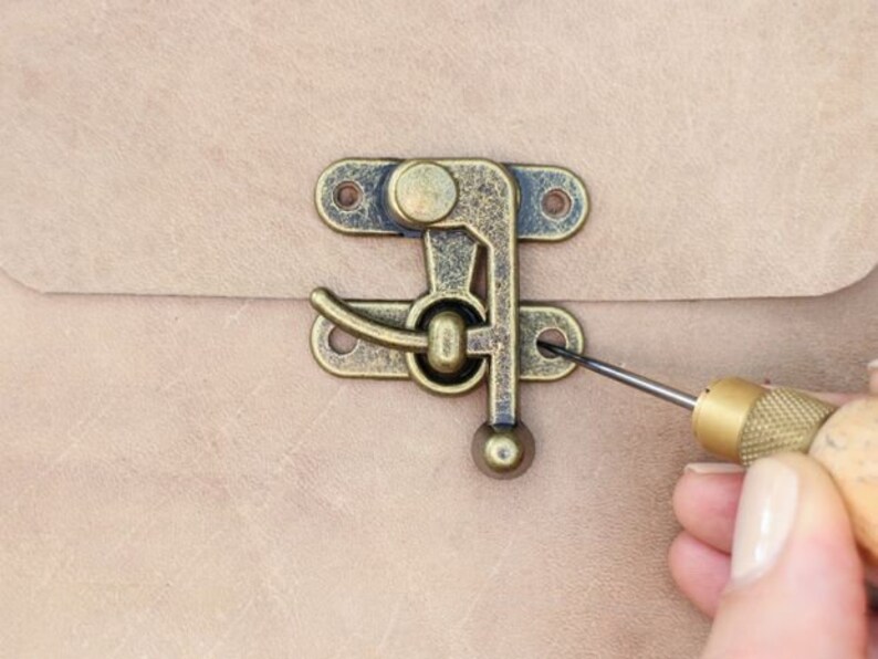 Pode incluir: Um close-up de uma trava de lat&atilde;o sendo fixada com uma pequena chave de fenda. A trava tem um design decorativo e est&aacute; presa a uma superf&iacute;cie de couro marrom claro.