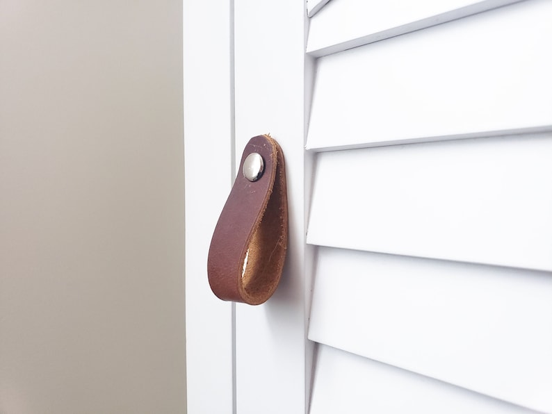 May include: A brown leather drawer pull with a silver rivet is attached to a white cabinet door. The pull is a loop shape, and the cabinet door has a shutter design. The background is a neutral colour.