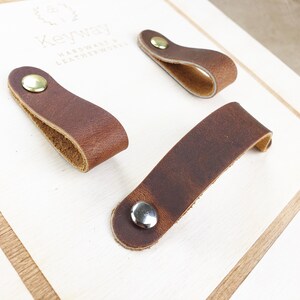 May include: Three brown leather drawer pulls with metal rivets are displayed on a light-coloured wooden surface. The leather has a distressed look, and the rivets are silver and gold-toned. The pulls are in different shapes.