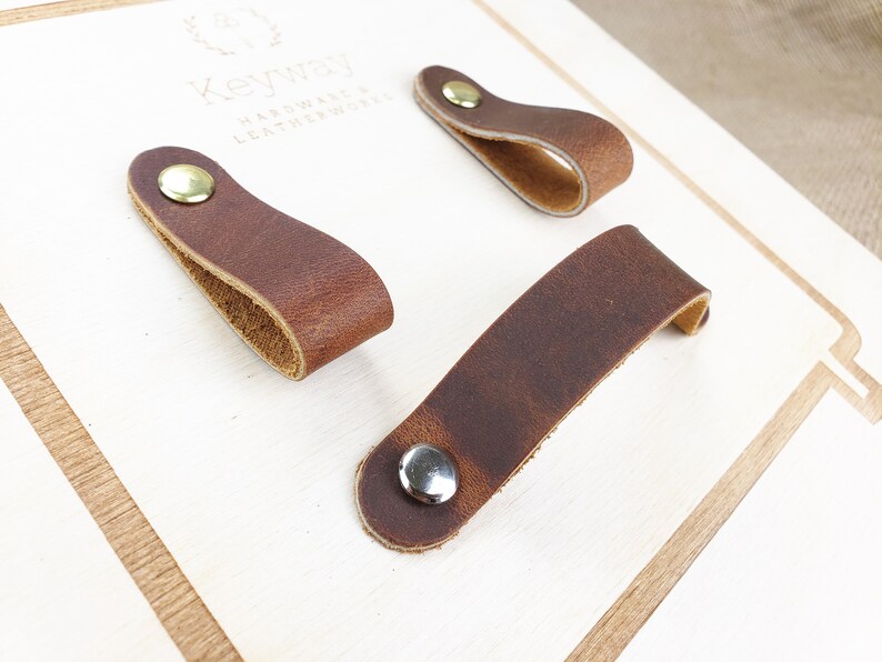 May include: Three brown leather drawer pulls with metal rivets are displayed on a light-coloured wooden surface. The leather has a distressed look, and the rivets are silver and gold-toned. The pulls are in different shapes.