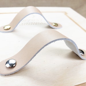 Könnte beinhalten: Zwei beigefarbene Leder-Schrankgriffe mit silbernen Metall-Akzenten. Die Griffe sind gebogen und haben ein schlichtes Design.