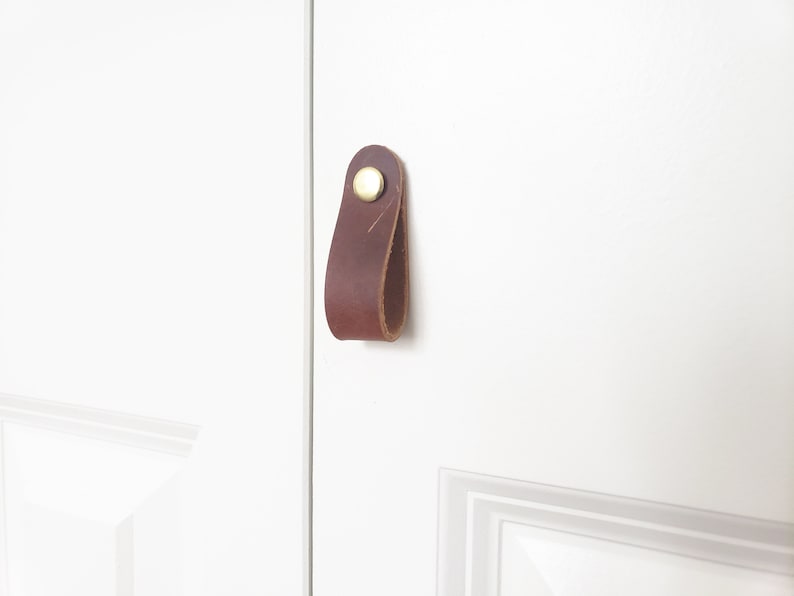 May include: A brown leather cabinet pull with a brass rivet. The pull is attached to a white door. This simple, modern hardware adds a touch of rustic charm to any cabinet or drawer.