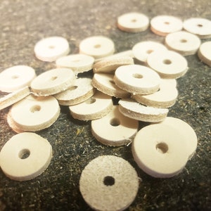 May include: A pile of off-white leather washers with a hole in the center. The washers are arranged in a random pattern on a brown surface.