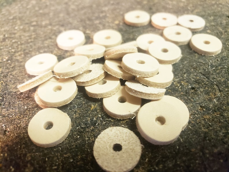 May include: A pile of off-white leather washers with a hole in the center. The washers are arranged in a random pattern on a brown surface.