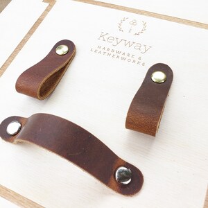 May include: Three brown leather cabinet pulls with metal rivets. One is a long, curved handle, and the other two are smaller, loop-shaped pulls. The pulls are on a light-coloured wooden surface. The text "Keyway Hardware & Leatherworks" is visible.