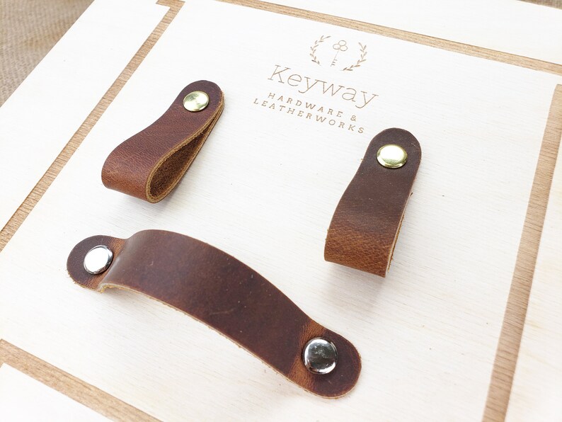 May include: Three brown leather cabinet pulls with metal rivets. One is a long, curved handle, and the other two are smaller, loop-shaped pulls. The pulls are on a light-coloured wooden surface. The text "Keyway Hardware & Leatherworks" is visible.