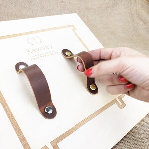 May include: Two brown leather drawer pulls with brass and silver fixings are shown on a light wood surface. One pull is being held by a hand with red nail varnish. The brand name "Keyway Hardware & Leatherworks" is visible.