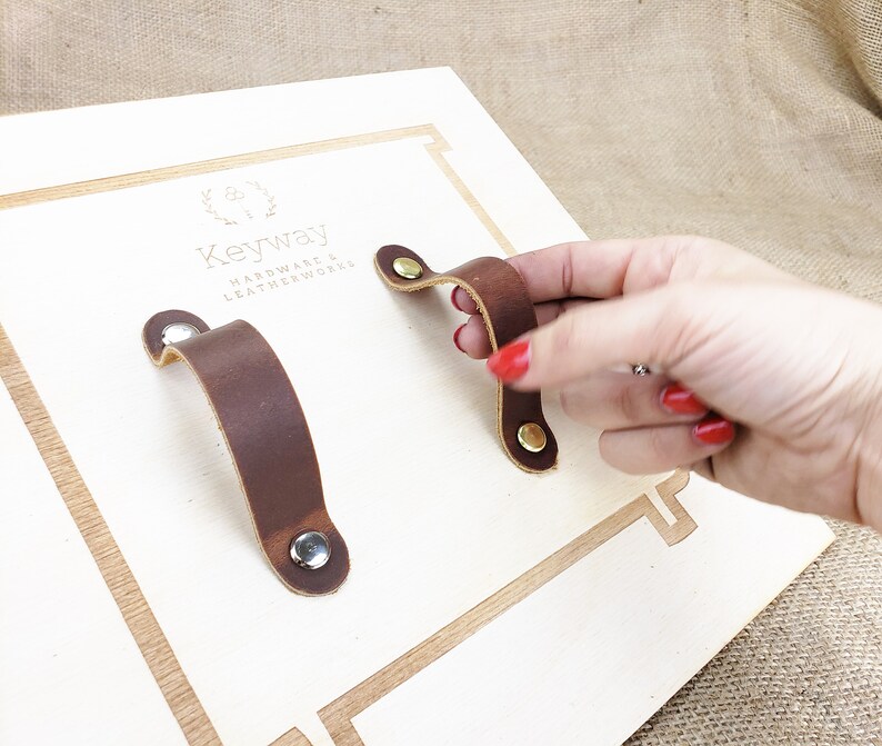 May include: Two brown leather drawer pulls with brass and silver fixings are shown on a light wood surface. One pull is being held by a hand with red nail varnish. The brand name "Keyway Hardware & Leatherworks" is visible.