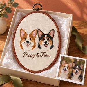 Op de afbeelding: Een ovale borduurring met een geborduurd portret van twee Corgi-honden, een bruin-witte en een zwart-bruin-witte, met de tekst Poppy & Finn eronder geborduurd. De ring wordt gepresenteerd in een geschenkdoos met een bijpassende foto.