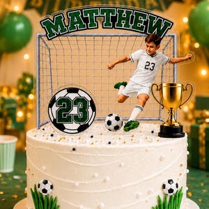May include: A white frosted cake topped with a soccer-themed acrylic topper featuring a soccer goal, a player in a white jersey with the number 23, a soccer ball, a gold trophy, and the name Matthew. The cake has small soccer ball accents.