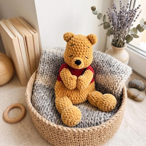 May include: A plush, golden-yellow crocheted bear toy with a dark red shirt and a small black nose. The bear is seated inside a round, woven jute basket lined with a thick, chunky-knit grey and white blanket, showcasing a soft, textured yarn finish.