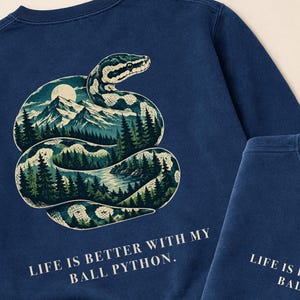 May include: A navy blue crewneck sweatshirt featuring a graphic of a coiled ball python with a landscape of mountains, pine trees, and a moon inside its body. The text "LIFE IS BETTER WITH MY BALL PYTHON." is printed on the front and back.