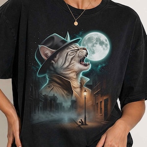 May include: A black short-sleeved t-shirt featuring a graphic print of a cat wearing a fedora and trench coat. The cat is depicted in a foggy, dark city street scene at night under a full moon, with a street lamp illuminating the cobblestone path.