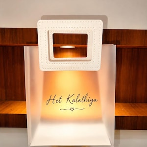 Op de afbeelding: Een doorschijnende witte rechthoekige tas met een stijf wit vierkant handvat met stikseldetails. De voorkant van de tas toont de zwarte tekst "Het Kalathiya" boven een klein decoratief hartsymbool, van binnenuit verlicht.