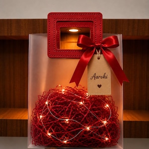 May include: A translucent gift bag with a red square handle and a red satin ribbon bow. A beige rectangular gift tag hangs from the bow with the name Aarohi and a heart symbol printed on it. The bag is filled with red shredded paper and fairy lights.