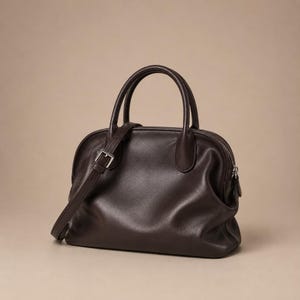 Puede incluir: Un bolso de mano color marrón oscuro con la parte superior redondeada, dos asas cortas y una correa ajustable para el hombro con una hebilla de metal plateado. El bolso tiene un acabado liso y mate con un cierre de cremallera en la parte superior.