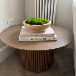 Könnte beinhalten: Ein runder Couchtisch aus Holz mit geriffeltem Sockel, darauf zwei gestapelte Bücher mit den Titeln The Kinfolk Home und Live Beautiful sowie eine helle Holzschale, die mit leuchtend grünem Moos gefüllt ist. Der Tisch hat eine glatte Platte.