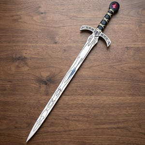 Könnte beinhalten: Ein silberfarbenes Schwert mit einer spitzen Klinge, die mit komplizierten schwarzen Linienmustern graviert ist. Der Griff besteht aus einem schwarzen Bereich mit goldenen Akzenten, einem silbernen Parier mit schwarzen Details und einem runden Knauf mit rotem Edelstein.