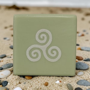 May include: A square, sage green bar of soap featuring a white triskelion symbol embossed in the centre. The soap has a smooth, matte finish and a flat surface, displaying a three-legged spiral design that radiates from a central point.