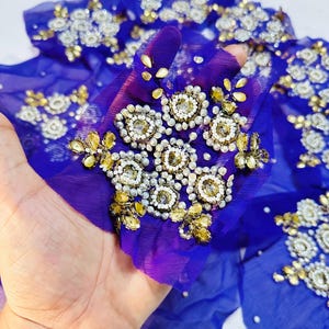May include: A piece of royal blue sheer fabric embellished with intricate floral-shaped clusters of clear white beads, small silver metallic beads, and faceted yellow-gold teardrop crystals arranged in a repeating pattern across the material.