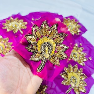 May include: A vibrant fuchsia fabric featuring multiple embroidered floral motifs. Each flower is crafted with gold sequins, metallic beads, and yellow thread, creating a textured, raised design across the bright pink material surface.