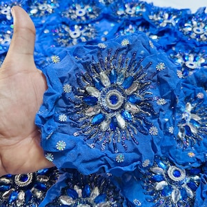 May include: A vibrant blue fabric featuring a repeating pattern of circular, ruffled rosettes. Each rosette is heavily embellished with a central blue stone, surrounded by clear teardrop-shaped crystals, silver metallic thread, and blue sequins.
