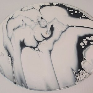May include: A white vinyl record with black and gray marbling and abstract patterns. The record has a circular shape and is painted with a swirling design.