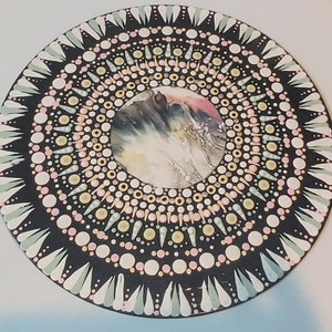 May include: A black circular wooden board with a mandala design painted in white, pink, and yellow. The center of the mandala features a swirling abstract design in black, white, and pink.