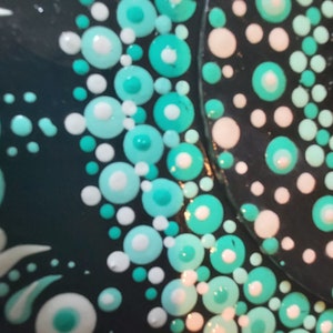 May include: Close-up of a black surface with a pattern of white and teal dots. The dots are arranged in a circular pattern, creating a mandala-like design.
