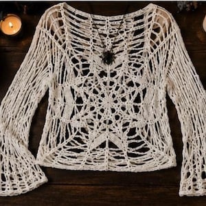 May include: A long-sleeved, off-white crochet top featuring an intricate, open-knit spiderweb pattern throughout the garment. The design includes a wide neckline and flared sleeves, constructed from a continuous yarn weave that creates a web-like texture.