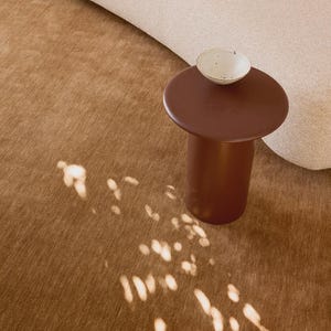 Op de afbeelding: Een donkerbruine cilindrische bijzettafel met een glad, rond bovenoppervlak. Op de tafel staat een kleine, gebroken witte keramische kom met een gespikkelde textuur. De tafel staat op een gestructureerd, lichtbruin oppervlak met lichtvlekken.