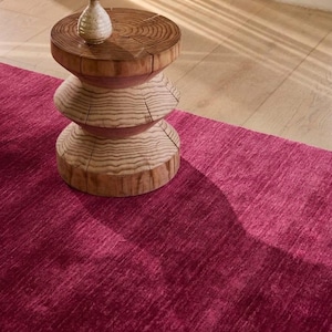 May include: A small, light-colored ceramic vase with a speckled finish sits atop a three-tiered, cylindrical wooden stool featuring visible grain patterns. The stool is placed on a deep magenta, textured area rug with a soft, plush appearance.