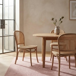 May include: A round light wood dining table paired with three chairs featuring curved wooden frames and woven cane backrests and seats. A ceramic vase with dried stems sits on the table surface, which is placed on a dusty pink area rug.