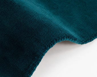 May include: A deep teal velvet fabric with a soft, dense pile and a subtle sheen. The material is folded, creating a curved edge that highlights the rich, saturated color and the plush, uniform texture of the textile surface.