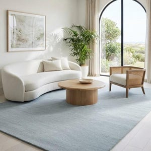 Puede incluir: Una alfombra rectangular grande de color azul claro sólido y apagado. La alfombra presenta una superficie texturizada de pelo corto y está colocada sobre un suelo de madera clara, sirviendo de base para un sofá blanco curvo y una mesa de centro.