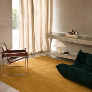 May include: A mustard yellow area rug positioned in a room with a brown leather and chrome chair, a dark green velvet floor cushion, beige linen curtains, and a concrete wall with built-in shelves holding a lamp and decorative objects.