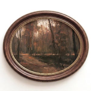 Könnte beinhalten: Ein ovales Wandkunstwerk mit einem dunklen, stimmungsvollen Waldlandschaftsgemälde in tiefen Braun-, Schwarz- und gedämpften Orangetönen. Das Kunstwerk ist in einen dicken, dunkelbraunen Holzrahmen mit einem dünnen, dekorativen goldfarbenen Innenrand gefasst.