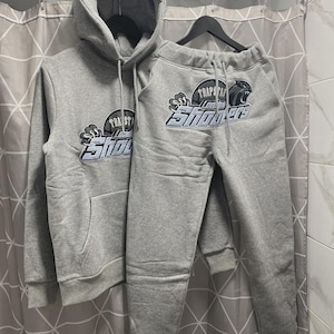 May include: A grey hooded sweatshirt and matching sweatpants set. Both pieces feature a dark grey and light blue embroidered graphic with the text Trapstar London Shooters. The hoodie has a front kangaroo pocket and the pants have a drawstring waist.