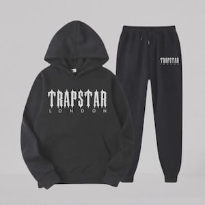 May include: A black hooded sweatshirt and matching sweatpants set. The hoodie features the white text TRAPSTAR LONDON in a gothic font on the chest. The sweatpants include a drawstring waist and the white TRAPSTAR LONDON logo on the upper left leg.